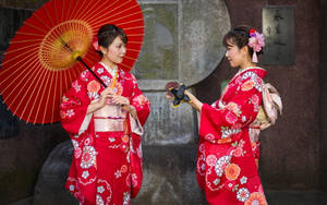 兩位身穿紅色花卉和服的女士在石祠附近手持陽傘和扇子。