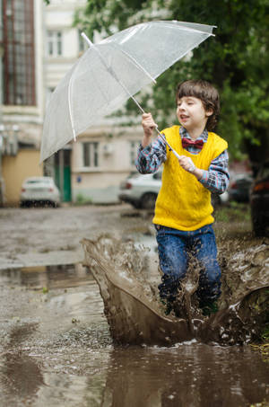 穿著黃色背心的孩子在雨天的街道上拿著透明雨傘跳進泥濘的水坑。