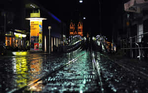 夜間城市街道上的雨，有電車軌道和遠處燈火通明的教堂。