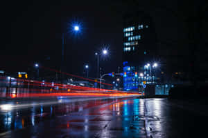 夜晚城市街道，潮濕的路面反射藍色燈光和紅色車燈條紋。 — MacBook Pro 14 壁紙
