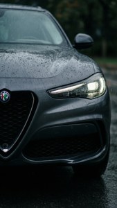 一輛深灰色愛快羅密歐汽車，引擎蓋上有雨滴，頭燈在夜間潮濕的森林中發光。