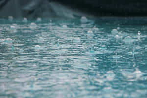 雨滴在藍綠色水面上泛起漣漪，後面有模糊的岩石。 — 雨天 壁紙