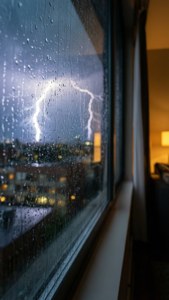 雨水順著窗戶流下，明亮的閃電在夜間的城市上空閃爍，溫暖的室內燈光可見