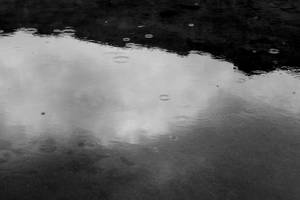 雨滴在倒映陰天天空的水坑上泛起漣漪的黑白圖像。