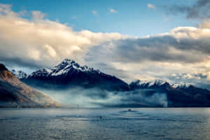 白雪覆蓋的山峰在霧氣瀰漫的湖泊後方升起，天空多雲而戲劇化，金色陽光穿透雲層。