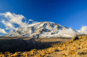 在清澈的藍天下，雲朵環繞著岩石山坡的雪山峰頂。