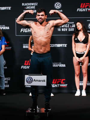 UFC拳手在稱重活動中於磅秤上展示肌肉，帶有品牌標誌和擂台女郎。 — 3:4 壁紙