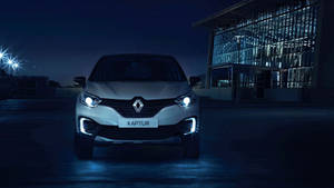 銀色雷諾Captur SUV在夜晚被車頭燈照亮，停在一棟現代玻璃建築前。