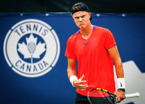 穿紅色襯衫的網球選手在比賽中表情猙獰，身後有Tennis Canada標誌。