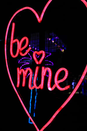 一個發光的粉紅色霓虹心形，上面寫着 'be mine'，背景是黑暗的城市夜景。 — 心形 壁紙