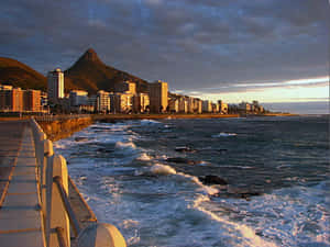 沿海城市在夕陽下，海浪拍打岩石，遠方有山。