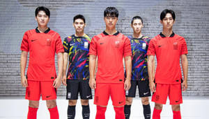 五名身穿紅色和圖案球衣的男足球員在磚牆前擺姿勢。 — 韓國 壁紙