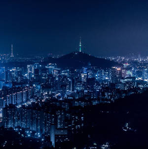 城市夜景，有發光的建築物和一座帶有發光塔樓的山。