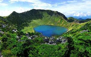 一個寧靜的藍綠色湖泊，位於山頂火山口中，周圍環繞著綠色植被和岩石地形。 — 韓國 壁紙