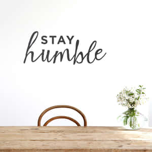 牆上寫有「STAY humble」文字，下方有木桌、椅子和白色花瓶。 — 語錄 壁紙