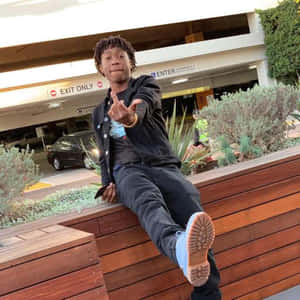 留著辮子的年輕男子坐在停車場木長椅上指著鏡頭。