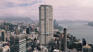 一座高大的玻璃摩天大樓矗立在城市建築中，靠近港口，遠處有山脈。 — 香港 壁紙
