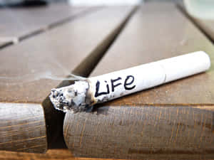 一支標有「Life」的燃燒香煙放在木質表面上，煙霧升起。 — 食草 壁紙