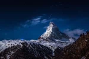 繁星點點的夜空下，白雪覆蓋的馬特洪峰，飄動的雲朵和下方的一盞藍光。