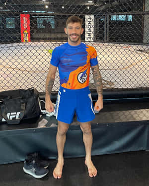 一位微笑的MMA戰士身穿藍色和橙色裝備，赤腳站在籠內。