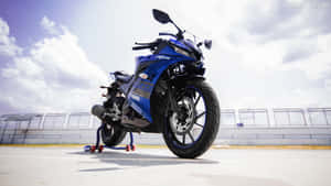 在多雲天空下的賽道上，藍色Yamaha R15運動電單車立於支架上。 — 低飽和度 壁紙