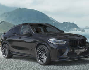 黑色Hamann BMW X6停泊在海岸碎石上，背後有海洋和山丘。 — 寶馬X6 M 壁紙