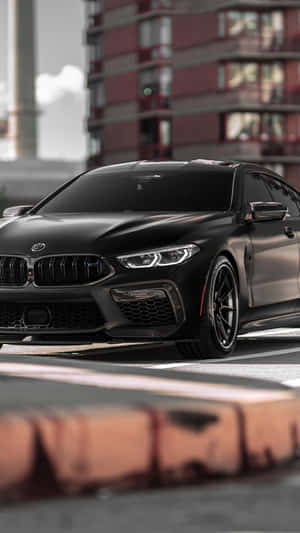 霧面黑色BMW M8 Coupe停喺城市街道上，前景係模糊嘅磚牆。