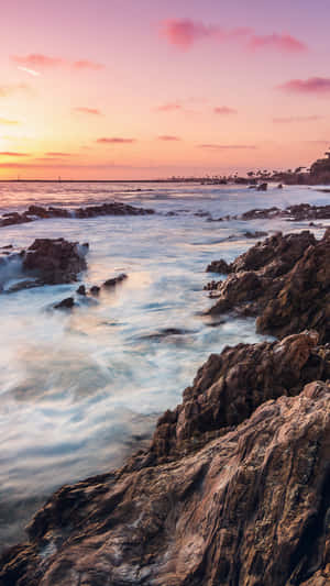 崎嶇的岩石在夕陽下與溫柔的海浪相遇，天空呈現粉紅色和橙色