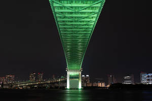 夜間一座綠色照明橋樑，城市燈光倒映在水面上。
