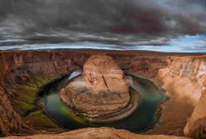 一條蜿蜒的河流在暴風雨的天空下，在深紅色的岩石峽谷中形成馬蹄形彎曲。 — 3:2 壁紙