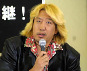 金髮男子穿著黑色皮褸和花卉圖案襯衫對著麥克風說話。
