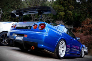 藍色日產Skyline R34 V-Spec，帶有大型後尾翼，停泊在秋樹附近的道路上。 — 深藍色 壁紙