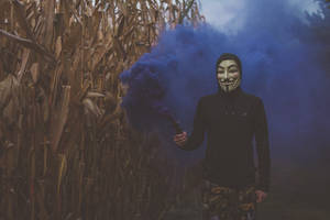 戴住蓋伊·福克斯面具嘅人喺粟米田釋放藍色煙霧。 — 鬼滅之刃面具 壁紙