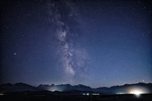 夜間積雪山峰上的銀河系與遠處燈光。 — 星星 壁紙