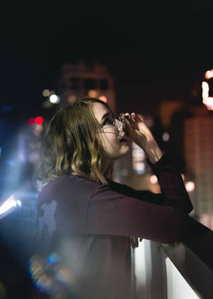女性在夜晚的城市陽台上從玻璃杯飲水，背景模糊燈光。