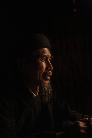 一位留著長鬍鬚的老人，在昏暗的燈光下若有所思地望向遠方。
