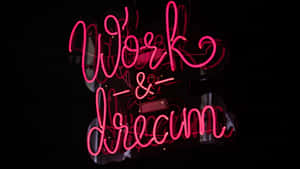 在深色反光表面上發光的粉紅色霓虹燈牌，上面寫著 Work & Dream。 — Studio Display 壁紙