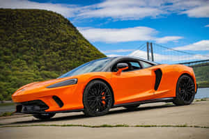 橙色McLaren跑車停泊在河邊，後面有橋和綠色山丘。