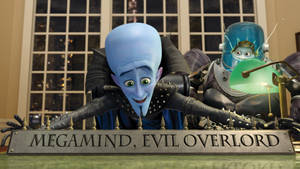 藍皮膚反派 Megamind 在標有「Evil Overlord」的桌子後咧嘴笑，旁邊有機械人助手。