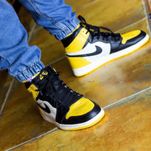 瓷磚地板上，穿著牛仔褲（褲腳摺起）的黃、黑、白色Nike Air Jordan 1運動鞋的人。