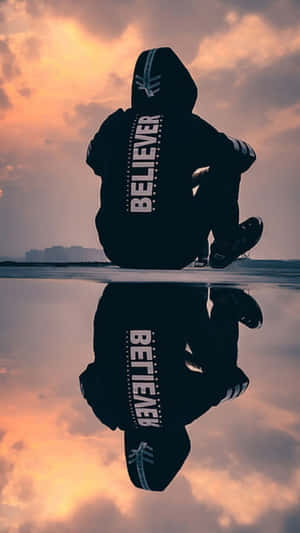 穿著印有「BELIEVER」文字的連帽衫的人的剪影，坐在水邊，帶有日落倒影。