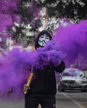 穿著黑色連帽衫和蓋伊·福克斯面具的人手持紫色煙霧信號彈在街上。 — 淨化面具 壁紙