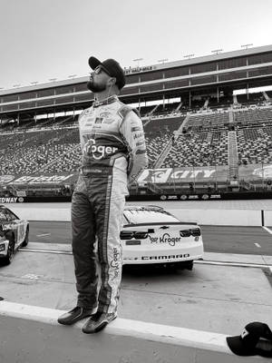 NASCAR車手身穿賽車服，在空曠的賽道上夕陽下站在賽車旁。 — 黑白 壁紙
