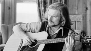 長髮男子室內彈奏木結他的黑白照片。