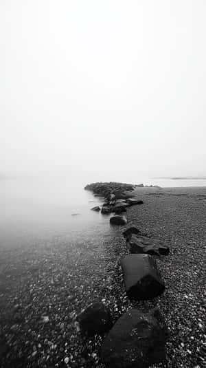 黑白海灘，岩石和鵝卵石在陰天天空下延伸至霧氣瀰漫的水面。 — 平靜 壁紙