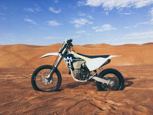 白色 Husqvarna 越野電單車停泊在沙漠沙丘上，藍天白雲。