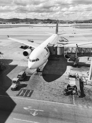 一架大型飛機停泊在機場閘口，地面服務車輛和遠山。 — 黑白 iPhone 壁紙