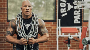 Dwayne Johnson 在磚牆健身房中用設備舉起沉重的鐵鍊。 — 岩石 壁紙