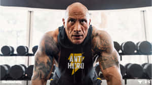 Dwayne Johnson 在健身房穿著帶有黃色閃電和文字的黑色連帽衫，周圍環繞著啞鈴。 — 岩石 壁紙