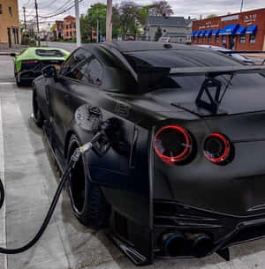 啞光黑色日產GT-R在加油站加油，後面有一輛綠色林寶堅尼。 — 汽車 壁紙
